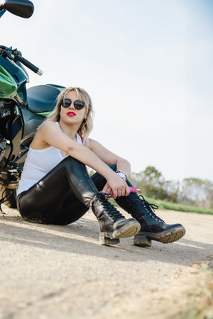 A woman in black pants and a white tank top sits casually on the ground beside a motorcycle. The image exudes a relaxed, adventurous vibe perfect for lifestyle and travel themes.の写真素材