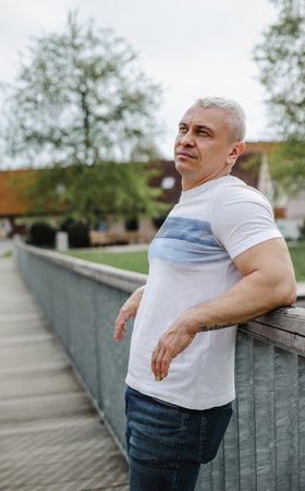 A man in a white shirt stands on a bridge, looking out over a park. He is in a contemplative mood,...
