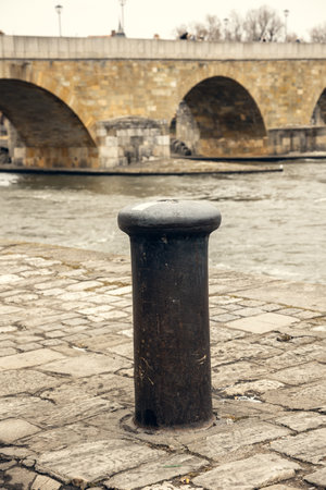 A worn mooring bollard on a historic cobblestone riverbank, set against the backdrop of an ancient stone bridge. Ideal for themes of history, travel, and urban heritageの写真素材