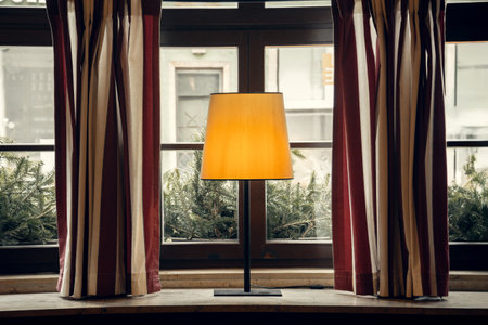 A warmly lit table lamp on a rustic windowsill, surrounded by striped curtains and greenery outside. A perfect composition for interior, decor, and cozy lifestyle projectsの写真素材