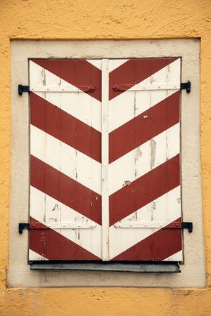 Aged wooden shutters with red and white diagonal stripes on a textured yellow wall. The peeling paint and vintage metal hinges add a rustic, historical characterの写真素材