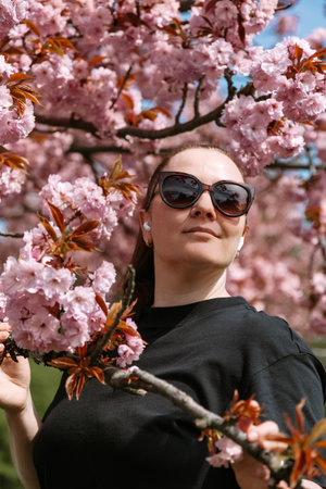 Portrait of a confident woman in sunglasses standing among pink cherry blossoms in spring. Great for seasonal concepts, empowerment, lifestyle design, and outdoor visuals.の写真素材