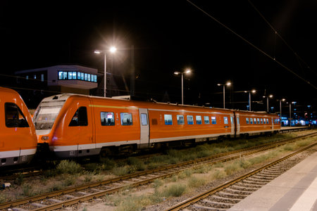 Orange passenger train parked on tracks at night station, illuminated by streetlights, representing travel, transport, and city commutingの写真素材