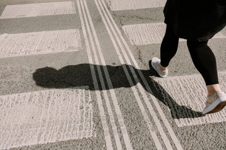 A woman confidently striding over a city crosswalk, captured from behind in bright sunlight. Perfect for themes of personal journey, direction, motivation, and lifestyle in urban settingsの写真素材
