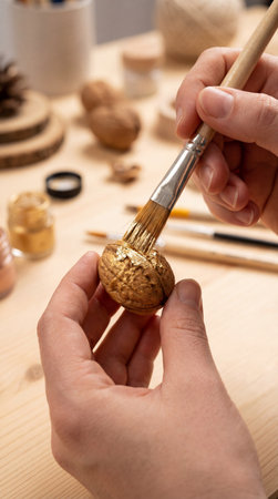 Close up hands painting walnut with gold paint brush. Creative DIY craft process on wooden table. Handmade holiday decoration preparation concept.の素材