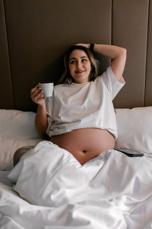 Pregnant woman sits bed holding cup smiling. Peaceful morning routine during pregnancy. Useful for...