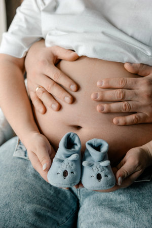 Close up of a pregnant woman's belly with her husband's hands resting on it. They are holding small...