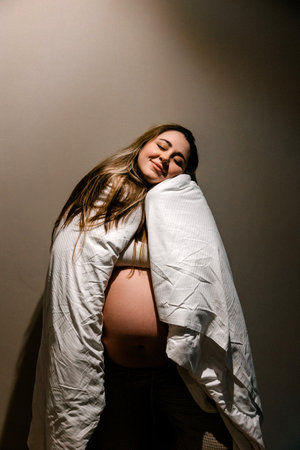 A pregnant woman smiles with eyes closed, wrapped in a white blanket. This image captures the...