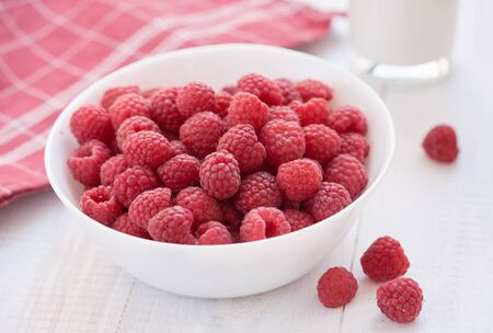 Red berries are in white bowl. In the background are red napkin and glass of milk.の写真素材