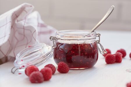 Glass jar with raspberry jam stands on table. Fresh raspberries are scattered nearby.