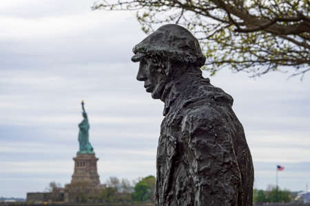 USA, New York, Ellis Island - May 2019: Statue, by Phillip Ratner, commemorating the millions of immigrants passing through Ellis Island, Statue of Liberty in back groundのeditorial素材