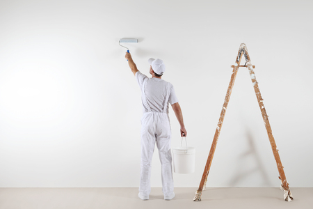 Rear view of painter man painting the wall, with paint roller and bucket, isolated on big empty space with wooden ladder.の写真素材
