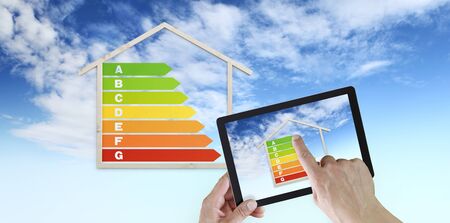 hand touch screen of digital tablet with house shape and energy efficiency chart symbol, isolated on blue sky background, green buildings and save energy eco sustainability.の写真素材