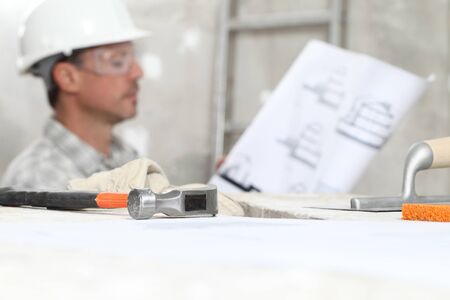 man work, professional construction worker looking at blueprints on interior building site, wear safety hard hat and protective glasses with plastering tools on scaffolding.の写真素材