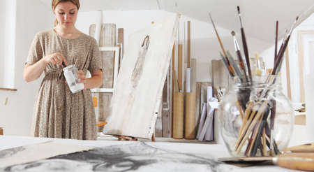 Indoor shot of young female artist in bright white studio while cleaning brushes wearing bohemian...