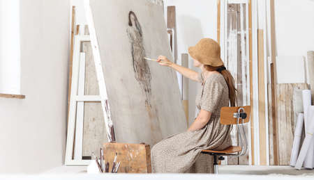 Indoor shot of young female artist sitting in front of the canvas and painting in bright white...