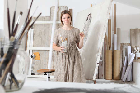 Indoor shot of young female artist looking at camera standing in front of the canvas holding painting tools in bright white studio wearing bohemian chic clothingの写真素材