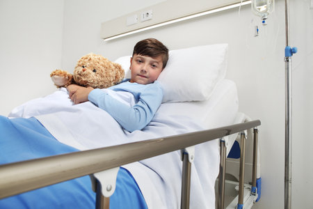 Portrait of cute child alone in hospital room lying in bed with teddy bear looking at camera with copy spaceの写真素材