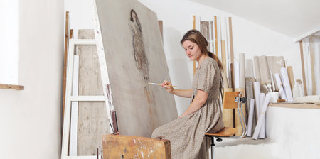 Indoor shot of young female artist standing in front of the canvas and looking at it holding...