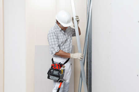 Man construction worker or plasterer holding drywall metal profiles near plasterboard white wall in building site. Wearing white hardhat, work gloves, safety glasses and tool belt.の写真素材
