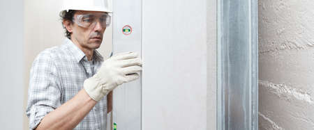 Man drywall worker or plasterer checking level of white plasterboard wall with bubble level at...