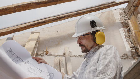 architect man at work, wearing helmet and headphones looking at blueprint, check the construction house project plan, in renovation building site background with the old wooden beams of the roofの写真素材