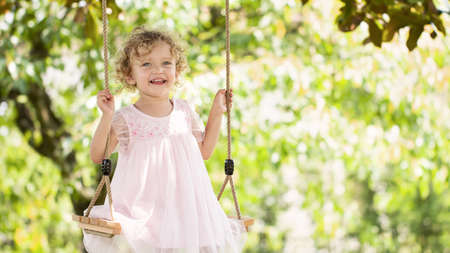 joyful smiling little girl swinging on the swing, child with blue eyes and curly blond hair plays in the green garden at home, concept of healthy growth and happy summerの写真素材