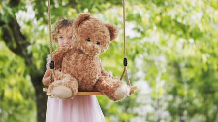 Spring and happy summer time. Joyful smiling little girl playing with teddy bear swinging on the swing. Child with blue eyes and curly blond hair in the green garden at home, concept of healthy growthの写真素材
