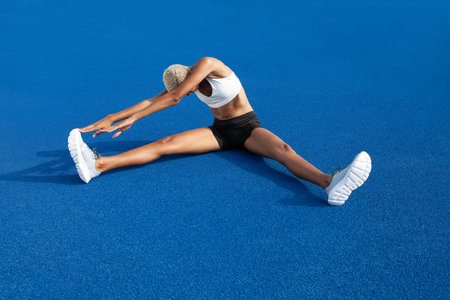 An African American female athlete engages in warm-up exercises, sitting on the blue track,...