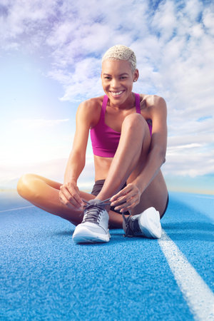An African American female athlete, smiling woman laces up her shoes sitting on the blue track,...