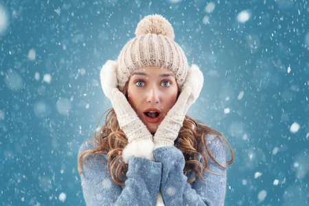 Amazed woman embracing the beauty of a winter snowfall. She is surprised at the falling snow during a white Christmas. Wearing cozy blue sweater, pom-pom beanie hat with ear warmers, scarf, and glovesの写真素材