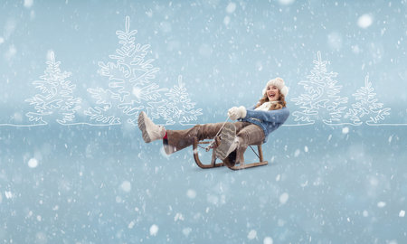 A cheerful and happy young woman sledding and enjoying the ride on sled, dressed in warm winter clothing, beanie hat, scarf, and gloves. A ski trip during the Christmas holidays in the winter seasonの写真素材