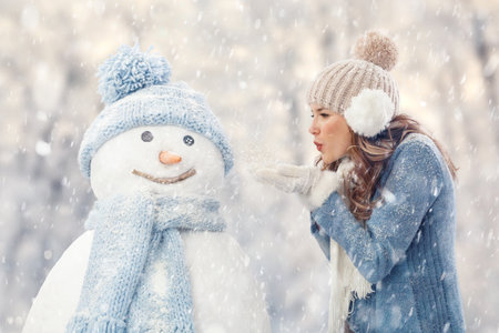 Happy young woman blowing snow from her hands onto snowman in warm winter clothing, outdoors in falling snow. Christmas holidays or winter season vacation conceptの写真素材