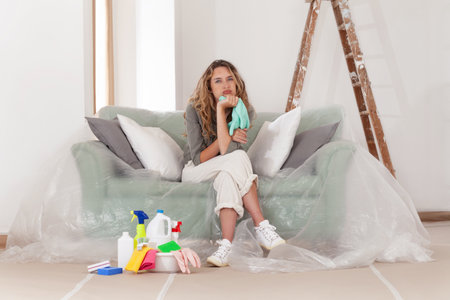 Tired, worried woman with household cleaning products beside her is sitting on the couch at home. She frustrated by the cleaning works during wall renovation and painting, typical spring cleaningの写真素材