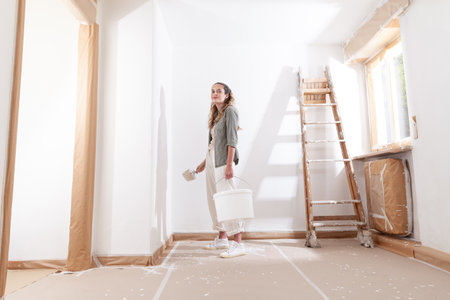 Happy woman painting home holding paintbrush and paint bucket in hand. Moment of DIY work for home decoration, renovating and refreshing the apartment in new rental or purchased property after movingの写真素材