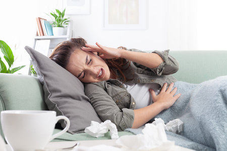 Young ill woman lies on the couch, holding her head and stomach in pain, suffering from a headache and abdominal discomfort, due to period cramps, flu, influenza or indigestionの写真素材