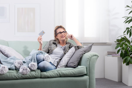 Smiling woman lying on a couch, wearing smart glasses and holding a credit card while interacting...