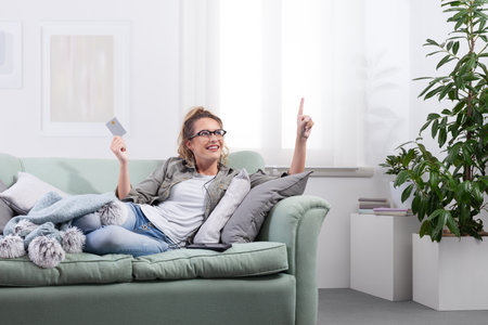 Smiling woman lying on a couch, wearing smart glasses and holding a credit card while pointing at a virtual screen. Concept of online shopping, digital payment, smart technology at homeの写真素材