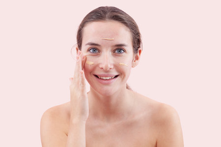 Smiling young woman with natural skin applying foundation cream on face. Visible makeup strokes....