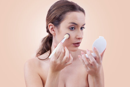 Young woman applying blush with a makeup brush while looking into a handheld mirror. Clean, minimal beauty routine concept. Isolated on beige background.の写真素材