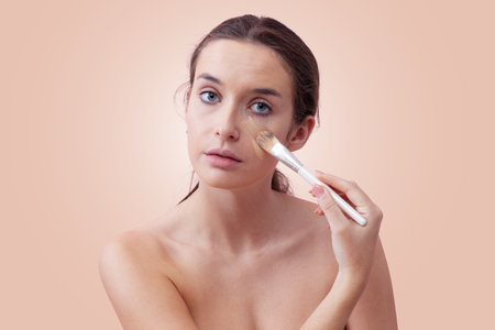 Young woman with natural skin applying foundation with a makeup brush, visible streak on cheek. Skincare and beauty routine concept. Isolated on beige background, clean and minimal lookの写真素材