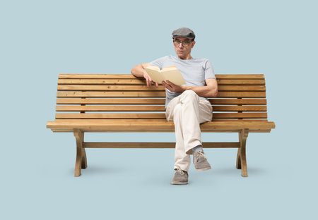 Mature man in his 50s sitting on a wooden bench and reading a book. Studio shot isolated on blue background. Wearing casual clothes, reading glasses, and a hat.の写真素材