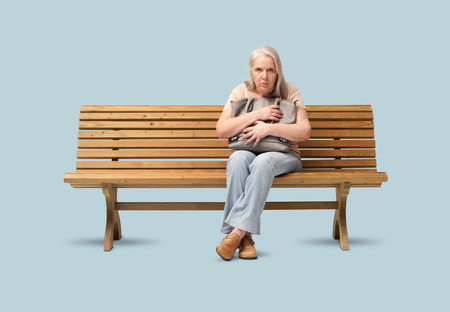 Scared mature woman sitting on a wooden bench, clutching her handbag tightly with a fearful expression. Concept of insecurity, anxiety, or fear of robbery. Isolated on blue backgroundの写真素材