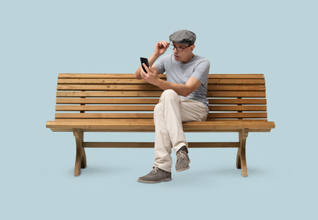 Mature man in his 50s sitting on a wooden bench and using a smartphone, studio shot isolated on blue background. Casual clothing, wearing reading glasses and a hatの写真素材