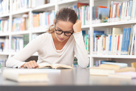 Young female university student studying in a library. She sitting at a desk and reading a book....