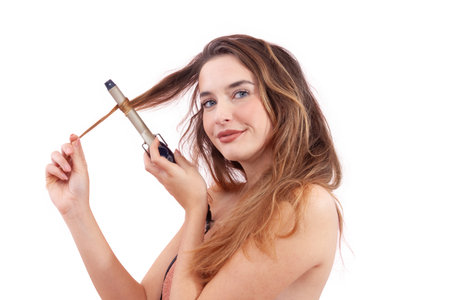 A cheerful young woman is styling her hair with a curling iron while smiling at the camera. Isolated on white background. Beauty, self-care, hair styling, and hair care themes conceptsの写真素材
