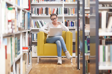 Young female university student studying in a library. She is sitting on an armchair, wearing...
