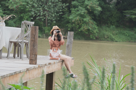 Young women are holding the camera sitting on the wooden bridge and river on background.And looking...