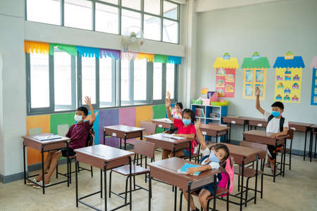 Group of school kids wear protective masks with teacher sitting in classroom and raising...