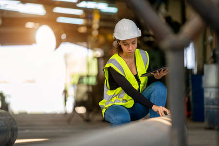 Young female in protective uniform inspecting industrial machine and taking necessary notes on digital tablet at plant.の写真素材